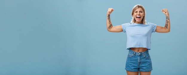 Free Photo strong women stand up and fight for rights portrait of happy entertained and cool young female athletic blonde with tattoos winking and smiling showing muscles and biceps over blue wall