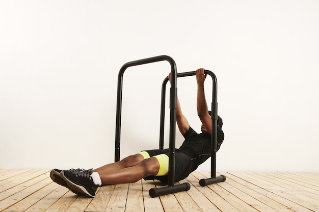 Strong young black male wearing black sports clothing with neon yellow half tights at the starting position for bodyweight rows on black mobile bars set on light wooden floor against white wall.