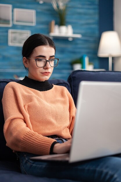 Student browsing online commerce information using laptop computer while sitting comfortable on sofa in living room. Woman studying marketing course using e-learning university platform