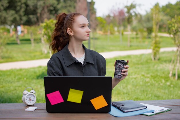 Student freelancer working in park holding coffee drinking looking up