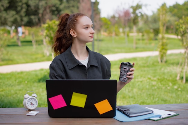 Student freelancer working in park holding coffee drinking looking up