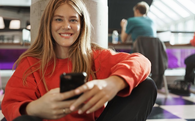 Free photo student lifestyle teenagers and urban life concept smiling happy attractive redhead girl sit on floor at cafe or coworking space texting friends using mobile phone playing smartphone