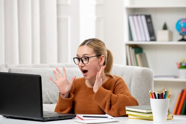 Student online young cute girl in glasses and orange sweater studying on computer shocked