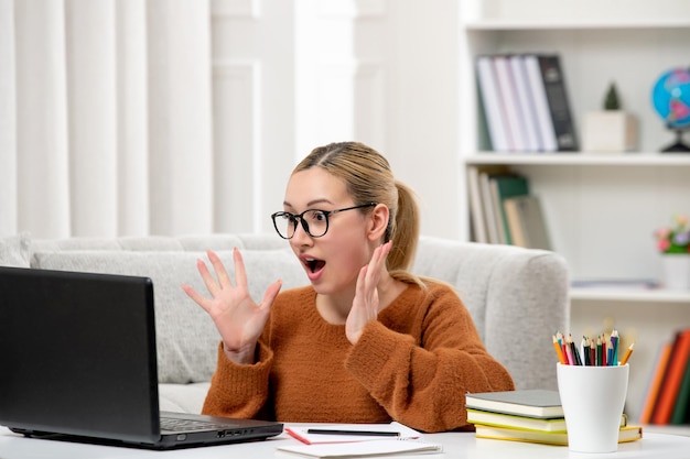 Student online young cute girl in glasses and orange sweater studying on computer shocked