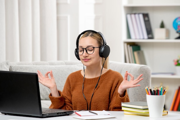 Student online young cute girl in glasses and orange sweater studying on computer showing zen sign