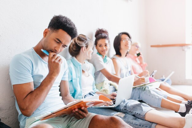 Students sitting on the floor with laptops and doing their homework together. Black boy with trendy hairstyle thinking about difficult task, holding pen in hand.