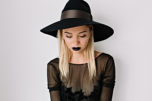 Studio close up portrait of pretty woman with dark lips is wearing hat and blouse is looking down over isolated background