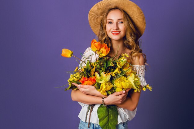 Studio  fashion portrait of pretty cute blonde woman in straw hat, white cotton  shirt sitting and holding bouquet of amazing spring  flowers. Wearing stylish retro  outfit .