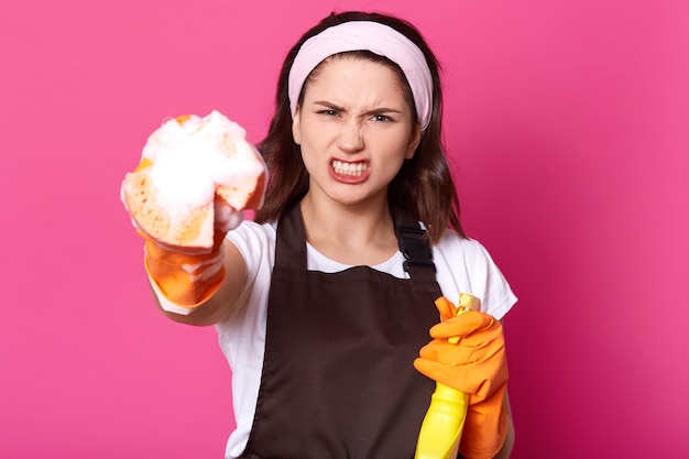 Free photo studio portrait of angry young caucasian woman dressed white t shirt and brown apron while cleaning up rooms, holding cleanser spray and sponge with foam in her hands, poses in photo studio.