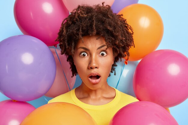 Studio portrait of astonished young woman posing surrounded by birthday colorful balloons