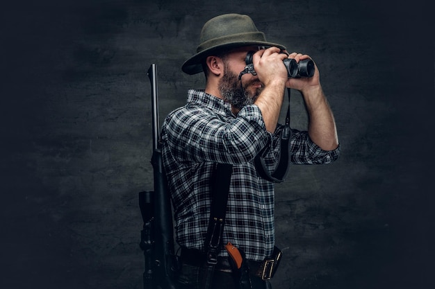 Studio portrait of bearded hunter male wearing a plaid fleece shirt looking through binoculars, holds a rifle.