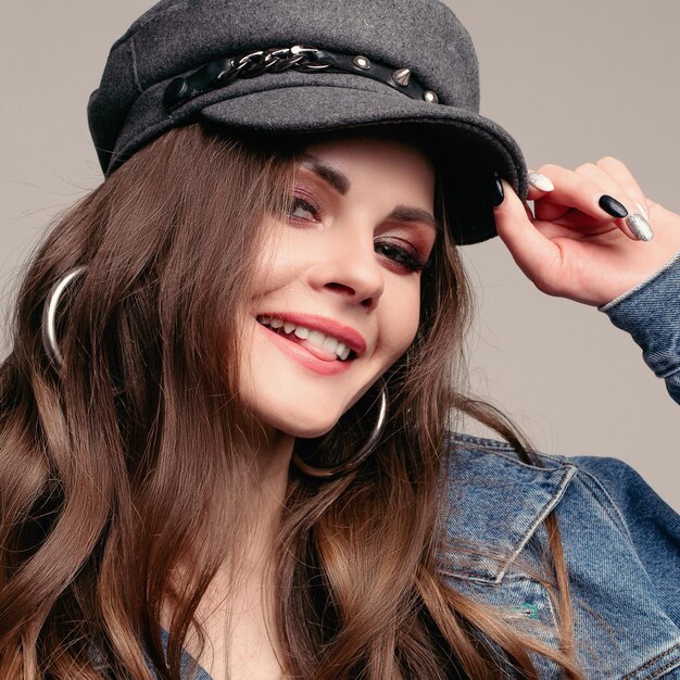 Studio portrait of beautiful brunette Caucasian girl with long wavy hair wearing denim jacket and stylish grey hat She is holding hat with manicured hand and smiling at camera Flirting with camera