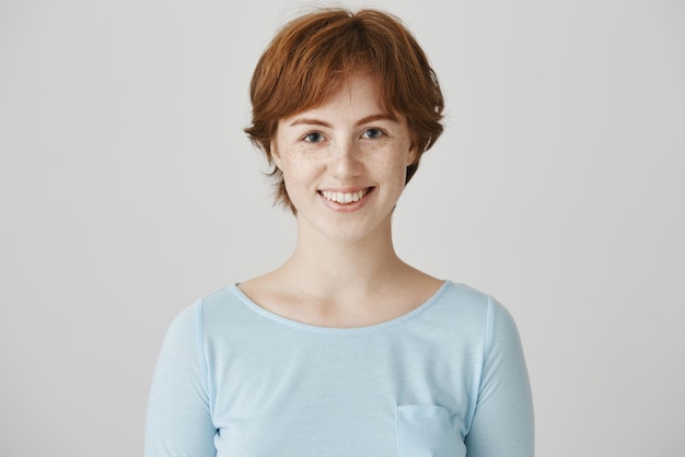 Studio portrait of charismatic attractive caucasian woman with natural red hair color and freckles smiling cheerfully while standing against gray background posing for advertisement