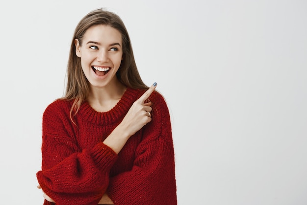 Studio portrait of charming satisfied young playful woman in stylish red loose sweater, smiling broadly, feeling excitement while pointing at upper right corner, standing 