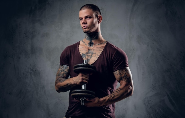 Free photo studio portrait of a man in a t shirt with tattoos on his arms  holds the dumbbell over grey artistic background.