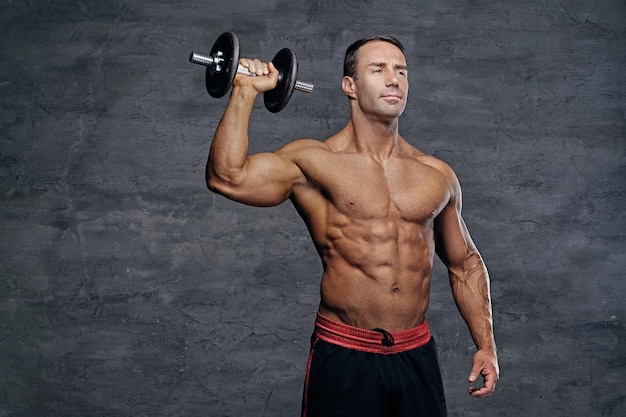 Studio portrait of shirtless muscular middle age male holds dumbbel isolated on grey background.