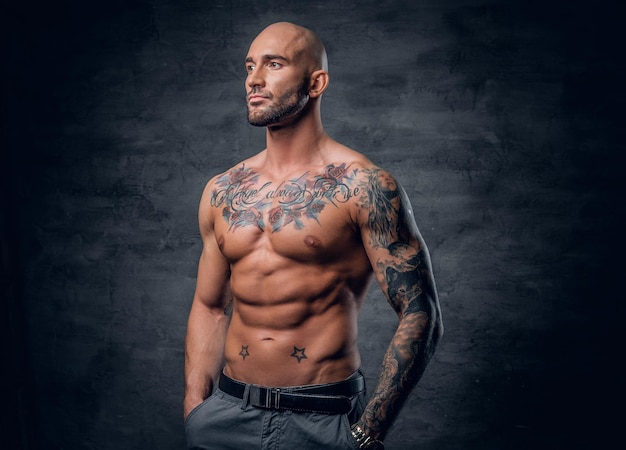 Studio portrait of shirtless shaved head, muscular male with tattoos on his torso over grey vignette background.