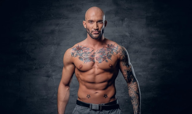 Free photo studio portrait of shirtless shaved head, muscular male with tattoos on his torso over grey vignette background.