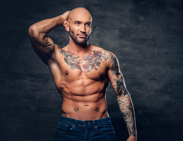 Studio portrait of shirtless shaved head, muscular male with tattoos on torso touch his head.