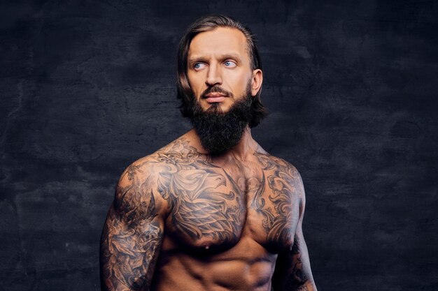 Studio portrait of shirtless, tattooed bearded male over dark grey vignette background.