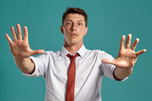 Studio portrait of a stately young guy in a classic white shirt and red tie acting like stopping someone and looking at the camera while posing over a blue background. Stylish haircut. Sincere emotion