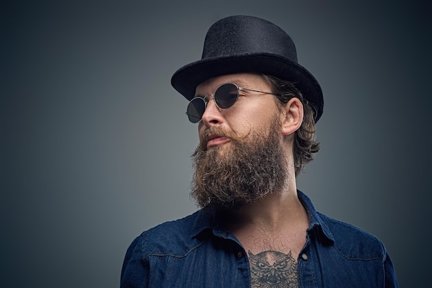 Studio portrait of stylish bearded male with a tattoo on his chest, dressed in a denim shirt, cylinder hat and sunglasses isolated on grey background.