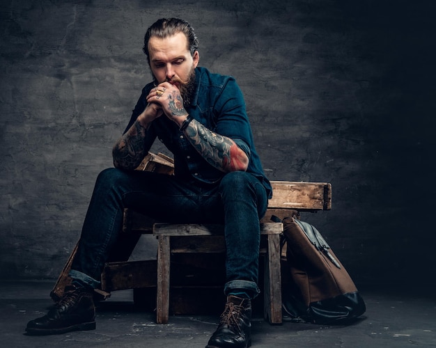 Free photo studio portrait of stylish bearded male with tattoos on arms, sits on wooden boxes over grey background in a studio.