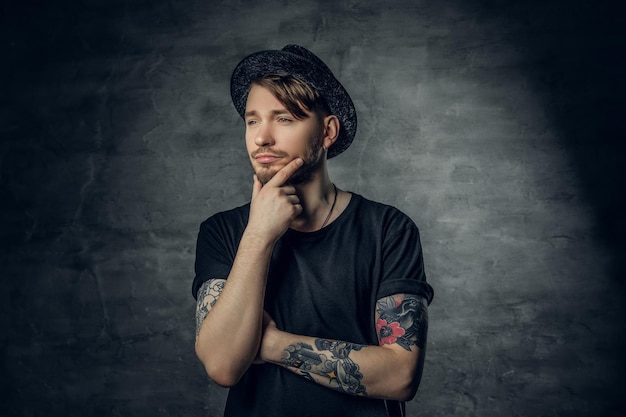 Free photo studio portrait of thoughtful male with crossed tattooed arms, dressed in a black t shirt and tweed hat.