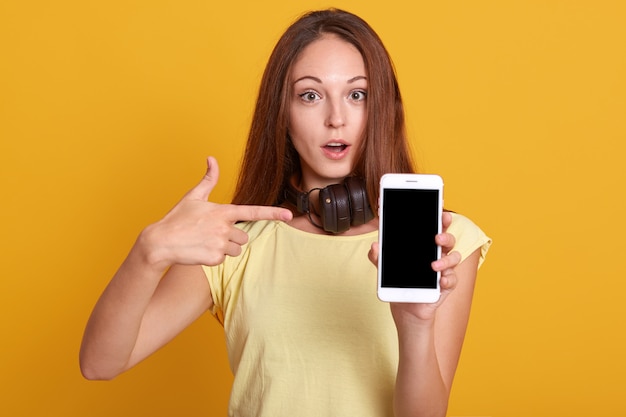 Studio sho of adorable woman showing phone with blank screen and pointing on it with her index finger