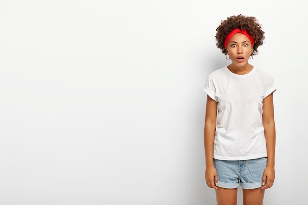 Studio shot of amazed hipster girl gazes at camera with opened mouth