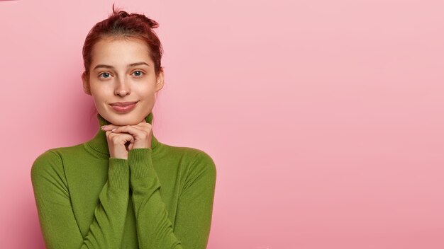 Studio shot of beautiful young Caucasian woman keeps palms under chin, gazes at camera with calm expression, wears green turtleneck, has natural beauty, isolated on pink wall, blank space aside