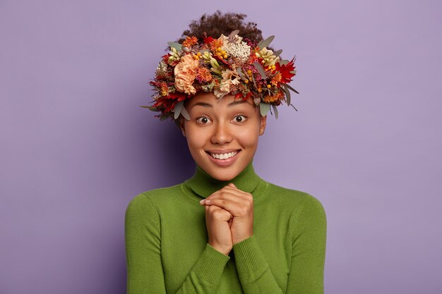 Free Photo studio shot of beautiful young lady smiles happily, keeps hands pressed together, looks hopefully at camera, wears autumnal handmade wreath, casual poloneck, models against purple background