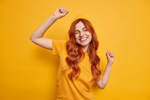 Studio shot of carefree redhead woman raises hand dances and smiles cheerfully has fun wears hat and casual t shirt 