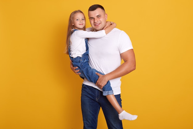 Studio shot of charming kid girl with her father, handsome man holding child in hands, wearing casual clothing