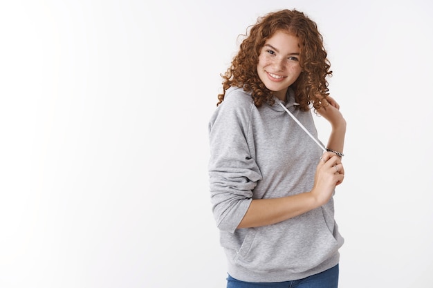 Studio shot cheeky cheerful redhead charismatic young girl wearing grey hoodie smiling silly flirting giggling playing hair wearing grey casual hoodie, standing white background amused lucky