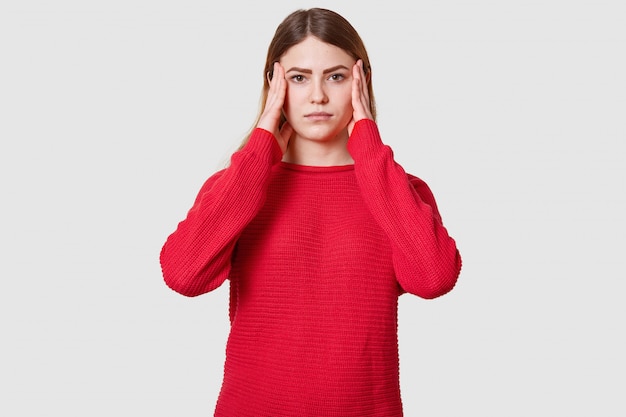 Studio shot of displeased female suffers from headache, dressed in red sweater, keeps hands on temples, has upset facial expression