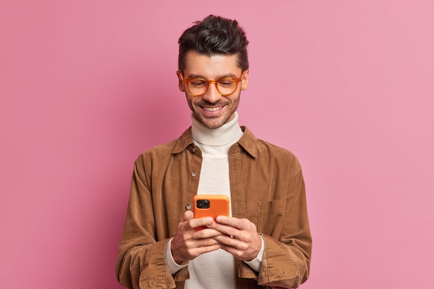 Free photo studio shot of european man blogger types text messages on smartphone smiles pleasantly