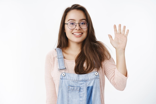 Studio shot of excited charming friendly girl meeting new people smiling broadly with amazement raising shoulders clumsy as waving hello raising palm in hi gesture gazing dreamy at camera