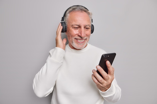 Studio shot of handsome senior man uses modern gadgets listens favorite music via headphones