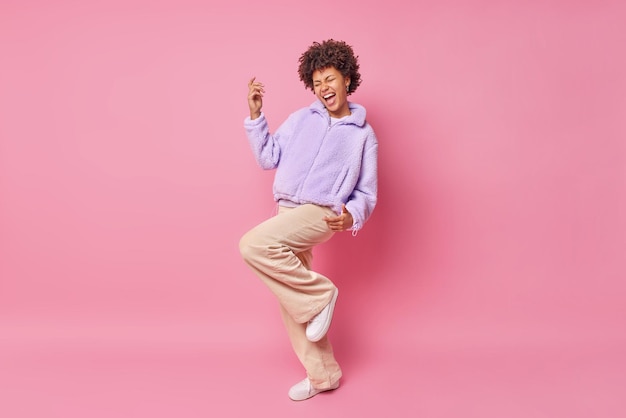 Free photo studio shot of happy curly haired woman makes triumph dance exclaims from joy wears purple fut jacket trousers and white sneakers isolated over pink wall