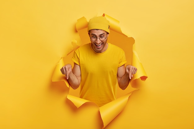 Studio shot of happy man with joyful facial expression, points down on floor, promots something, shows direction in bottom