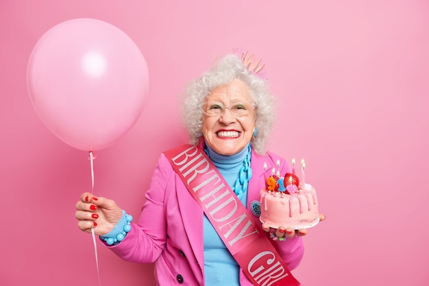 Studio shot of happy wrinkled female pensioner with bright makeup smiles toothily holds festive cake with burning candles has festive mood carries inflated balloon