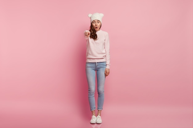 Studio shot of impressed young female has surprised expression, points directly at camera, being speechless