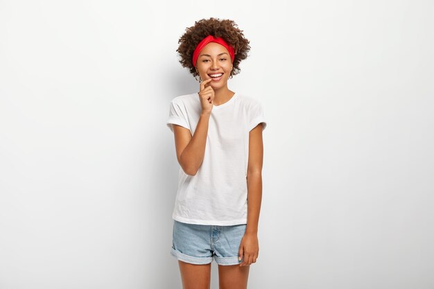 Studio shot of lovely curly haired woman enjoys pleasant moment, smiles gently, wears red headband, casual t shirt and denim shorts, isolated on white background