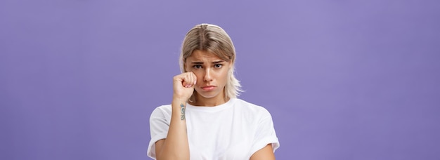 Free photo studio shot of offended sad and timid silly woman with blond hairstyle frowning looking from under forehead holding fist near eye as if whiping teardrop being upset over purple background