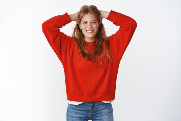 Studio shot of optimistic charming attractive young woman with cute freckles smiling joyfully with nice white smile holding hands on back of head in carefree chill pose wearing oversized red sweater