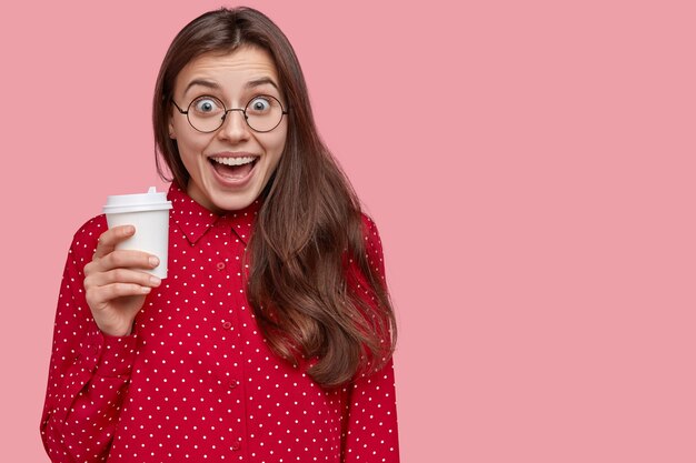 Studio shot of pleased young woman laughs positively, drinks takeaway coffee, has break after lectures