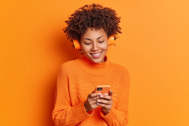 Studio shot of pretty dark skinned woman chooses song from playlist uses smartphone and wireless headphones