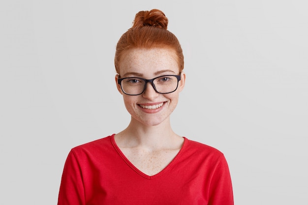 Free photo studio shot of red haired young smiling female wears square spectacles and red sweater, being in good mood after promotion at work, recieves award for diligent work and achievent great success