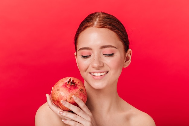 Free photo studio shot of relaxed girl with pomegranate. smiling ginger woman holding garnet on red background.